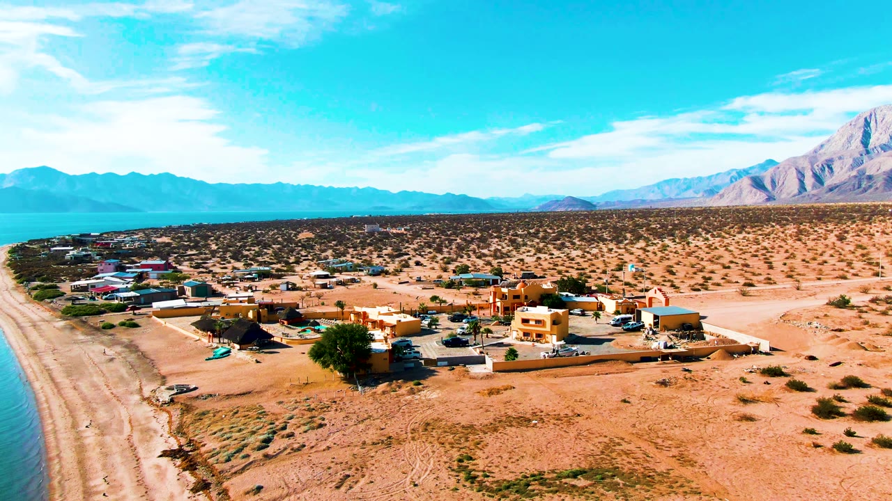 Aerial frame over Bahia De Los Angeles showing the village slightly closer, with a beach strip at left, orange and tan buildings near the center, open desert to the right, distant mountains, and no visible people.