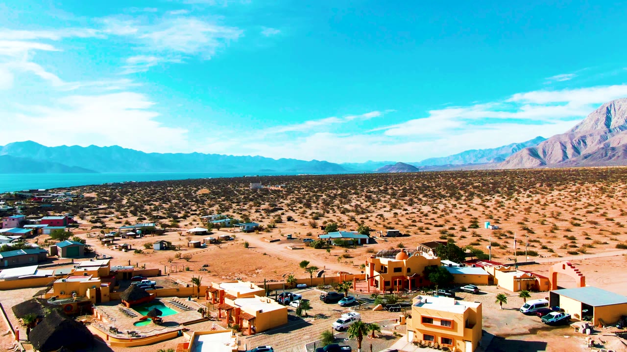 Opening aerial frame over Bahia De Los Angeles with tan buildings and parked vehicles in the foreground, desert scrub beyond, blue water and mountains on the horizon, and no visible people.