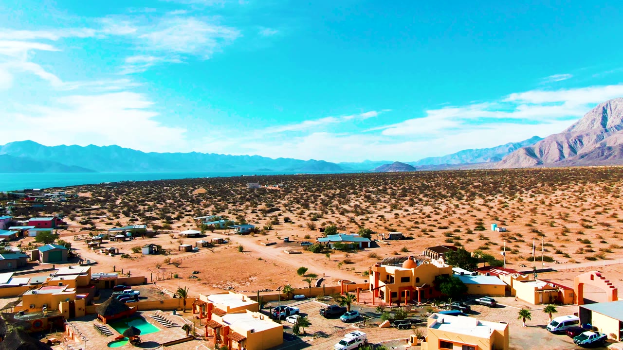 Second frame of Bahia De Los Angeles showing the elevated view shifted across low buildings, open sandy lots, parked vehicles, distant blue water, mountains, and no visible people.