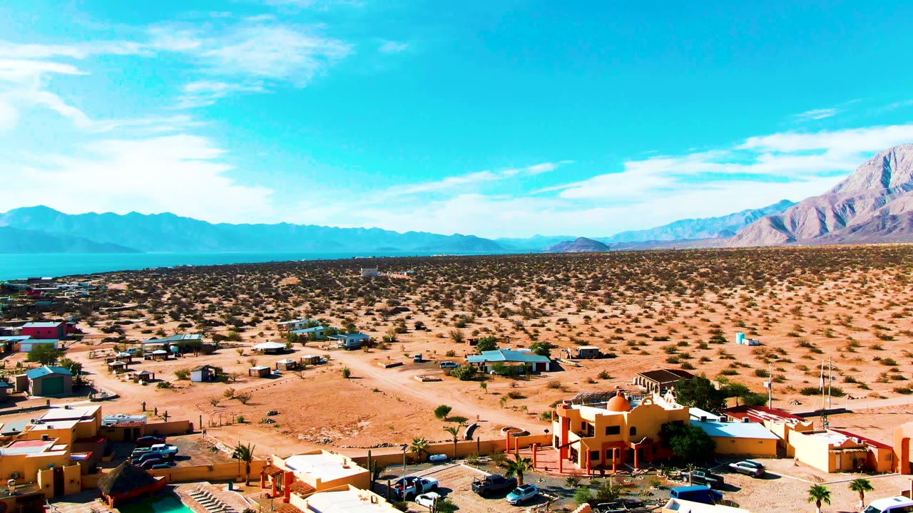 Third frame over Bahia De Los Angeles with warm-colored rooftops at the bottom edge, a dirt road and parked vehicles nearby, desert lots stretching to water and mountains, and no visible people.