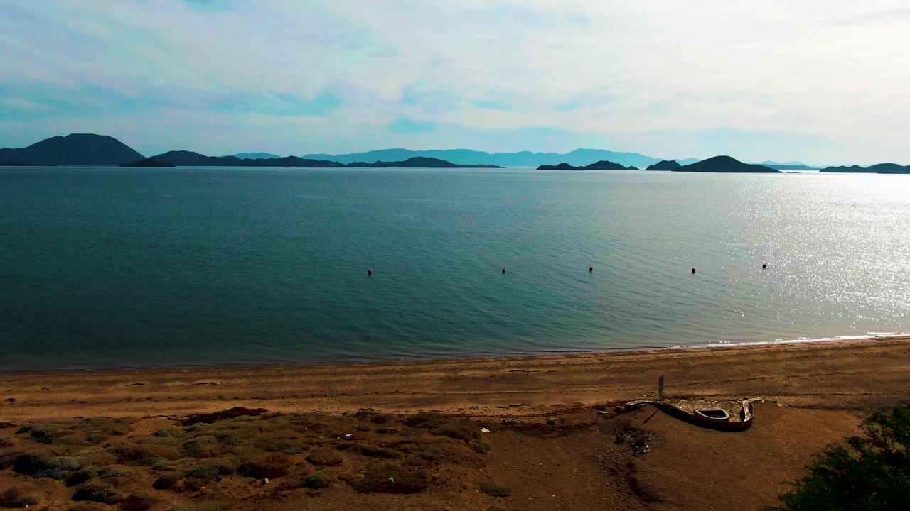 Wide view of Bahia De Los Angeles with dry sandy foreground, a narrow beach, calm blue water, distant island silhouettes, bright reflection on the right, and no visible people.