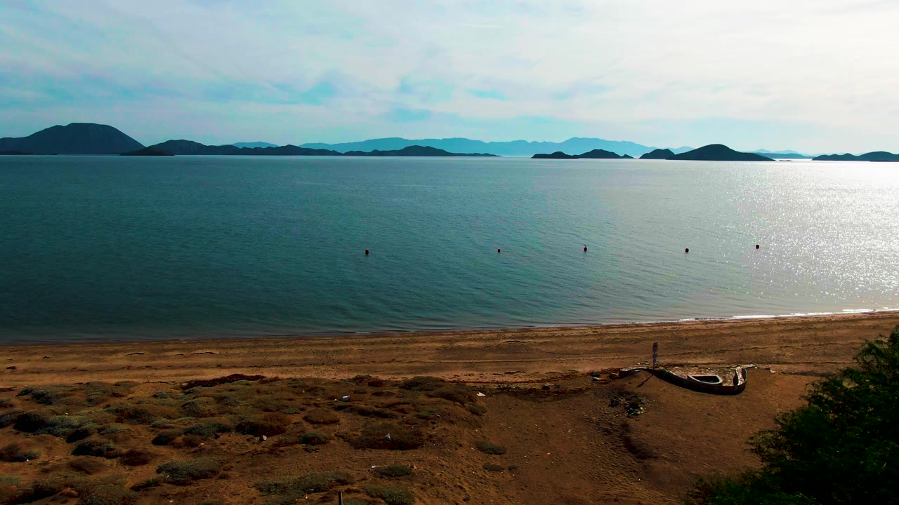 Bahia De Los Angeles shoreline with scrubby dry ground in front, calm blue ocean across the middle, small buoys offshore, hazy island ridges on the horizon, and no visible people.