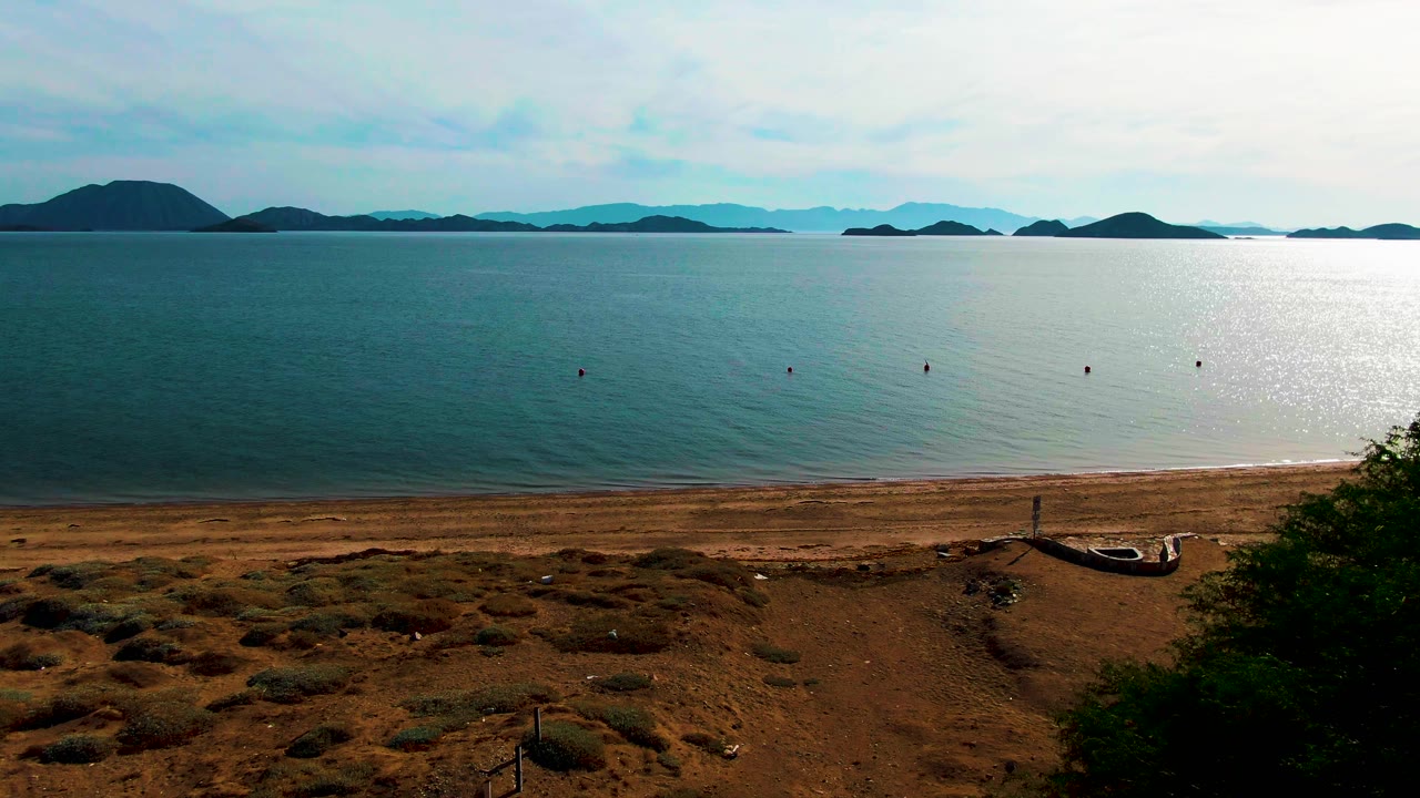 Rightward pan progress over Bahia De Los Angeles, showing more beach and dry scrub below, calm bay water with several small buoys, island silhouettes across the horizon, and no visible people.