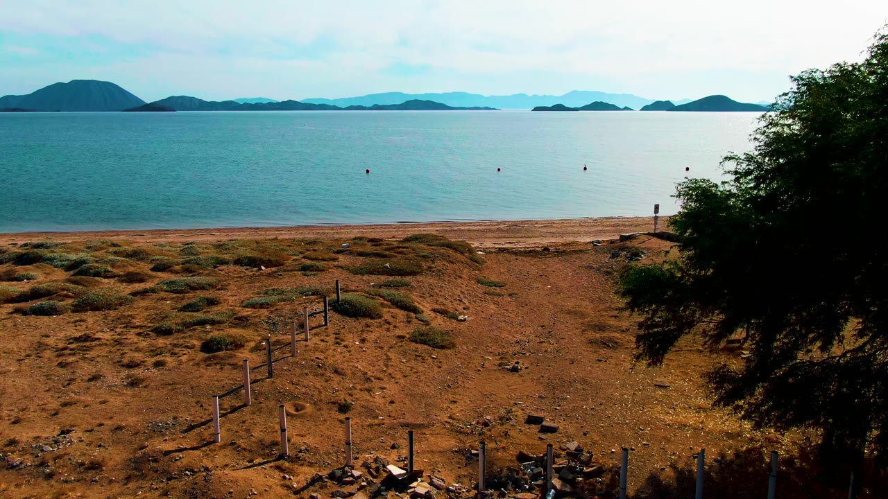 Wide coastal shore with dirt path, fence posts, calm water, and distant islands.