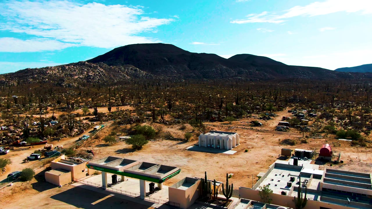 Aerial frame in Catavina BC Mexico showing low desert buildings at the bottom edge, sandy ground, scattered cacti, dirt roads, dark mountains, blue sky, and no visible people.