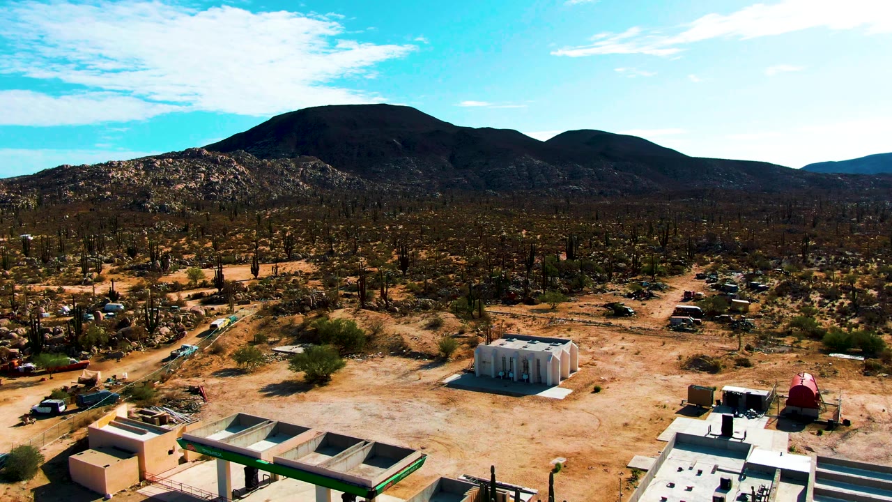 Catavina BC Mexico aerial frame with low buildings and a red-roofed structure near the bottom, parked vehicles along dirt tracks, cactus-covered desert, mountains, blue sky, and no visible people.
