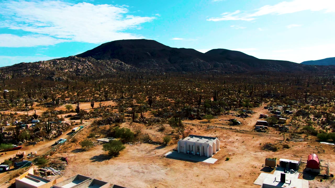Wide Catavina BC Mexico desert frame with buildings dropping toward the lower edge, dirt paths crossing sandy terrain, parked vehicles, many cacti, distant mountains, bright sky, and no visible people.