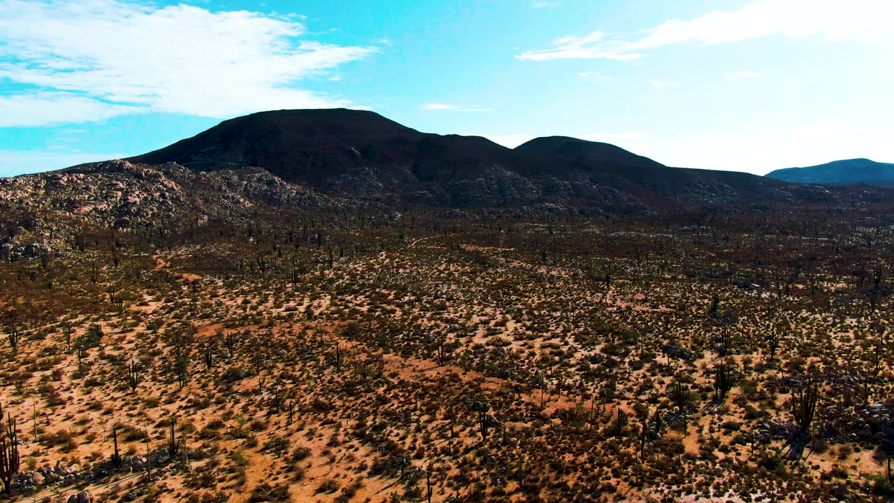 Aerial view over the Catavina desert in Baja California, Mexico, as the drone begins lateral movement; the dark volcanic mountain occupies the upper center, rust-orange desert floor with scattered cacti and scrub extends across the foreground, vivid blue sky above; no visible people.