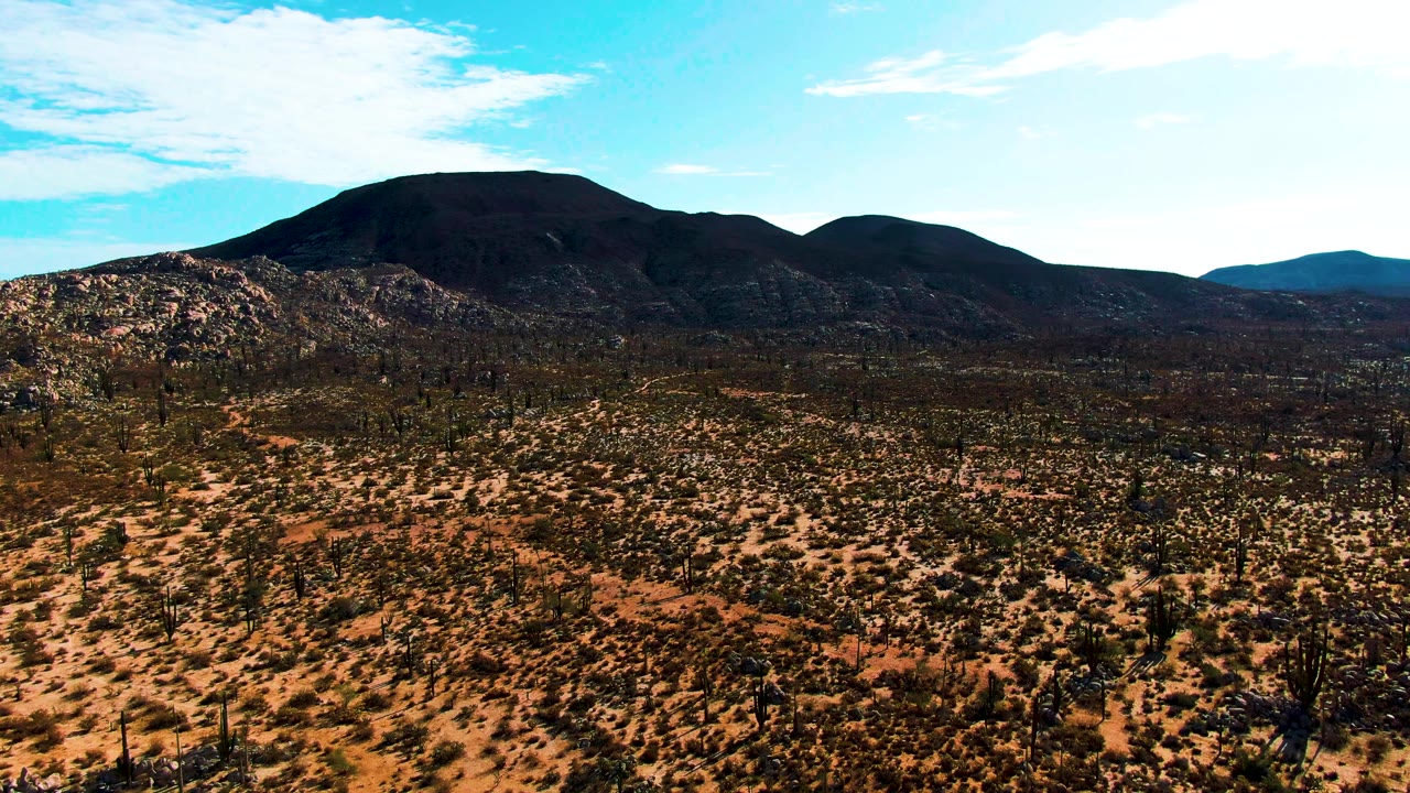 Aerial frame over the Catavina desert in Baja California, Mexico, with the dark volcanic mountain shifted slightly as the drone continues its lateral pass; rust-red arid terrain with scattered low cacti and desert scrub fills the foreground, vivid blue sky above; no visible people.