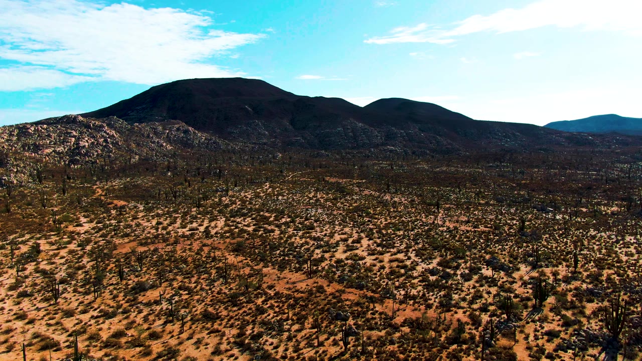 Aerial view over the Catavina desert in Baja California, Mexico, at mid-clip; the dark volcanic mountain remains prominent against the blue sky while rust-orange rocky terrain dotted with sparse low cacti and desert shrubs occupies the foreground; no visible people.