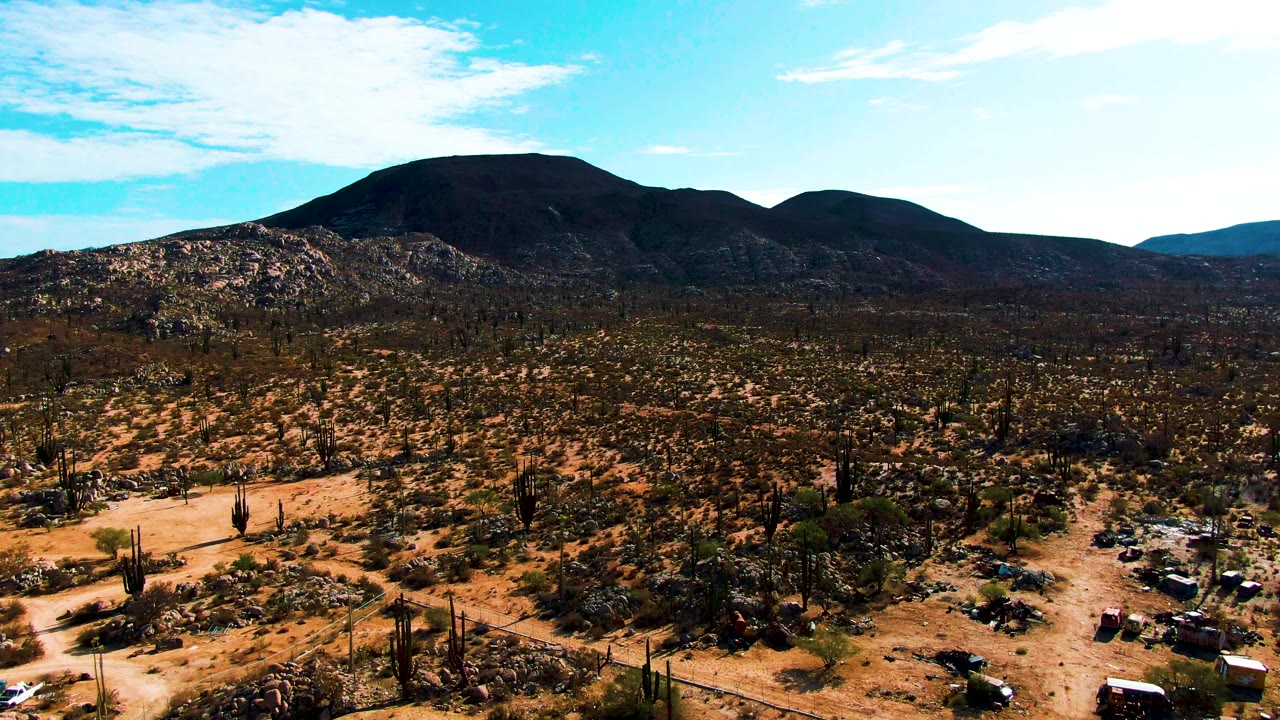 Aerial frame in Catavina BC Mexico with vehicles and campsite objects low in view, cactus-covered desert across the midground, dark rocky hills on the horizon, bright blue sky, and no visible people.