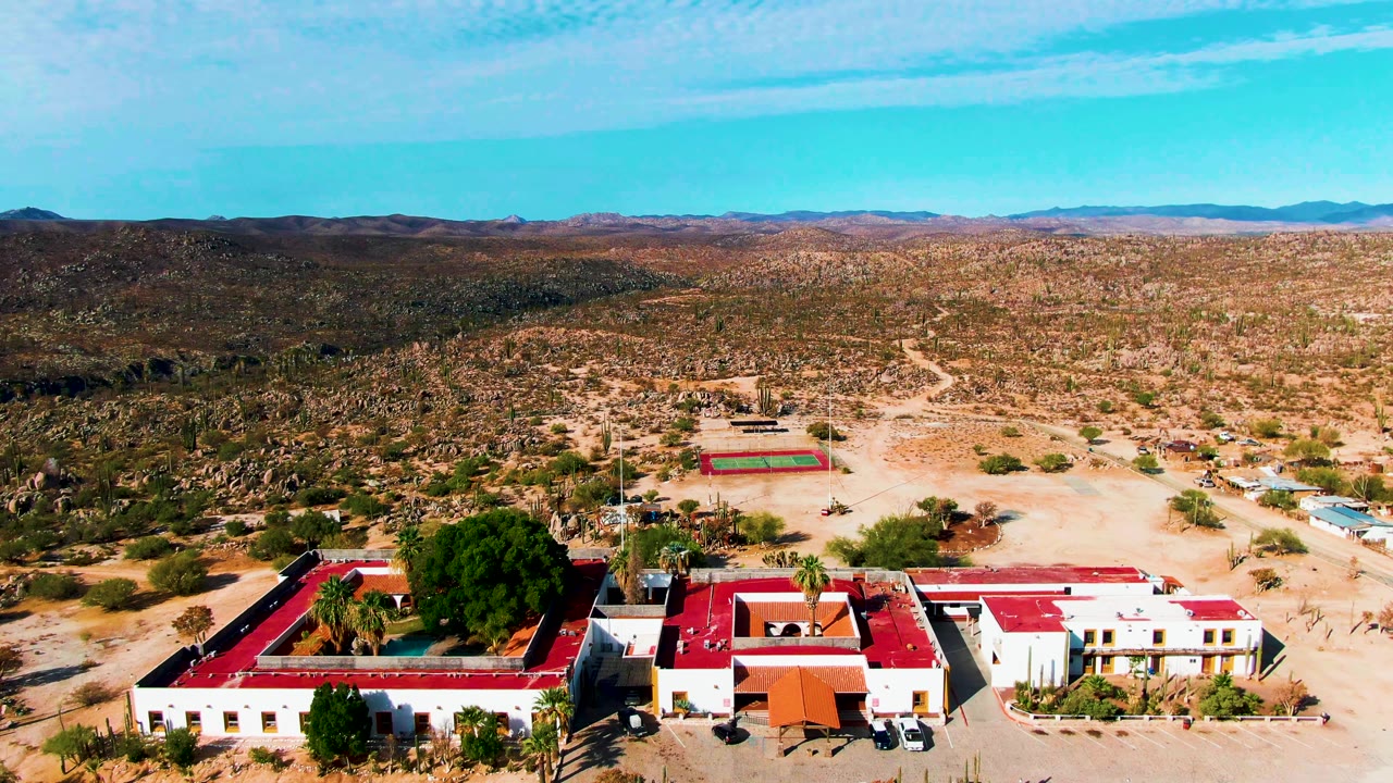 Aerial view over red-roofed white resort buildings, desert terrain, and a tennis court.