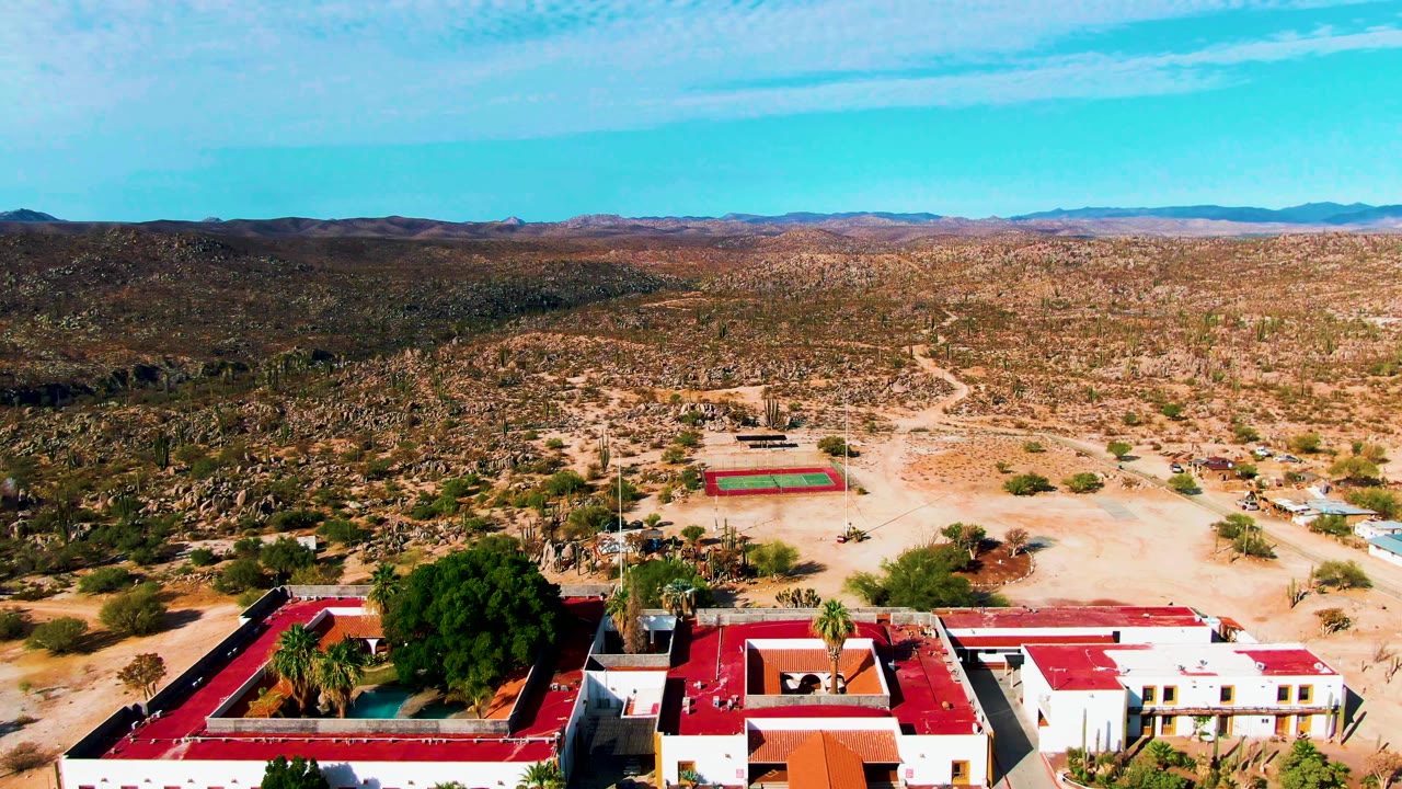 Catavina BC Mexico aerial view with the red-roofed resort lower in frame, a green tennis court with red border in the midground, scrubby desert, mountains, and no visible people.