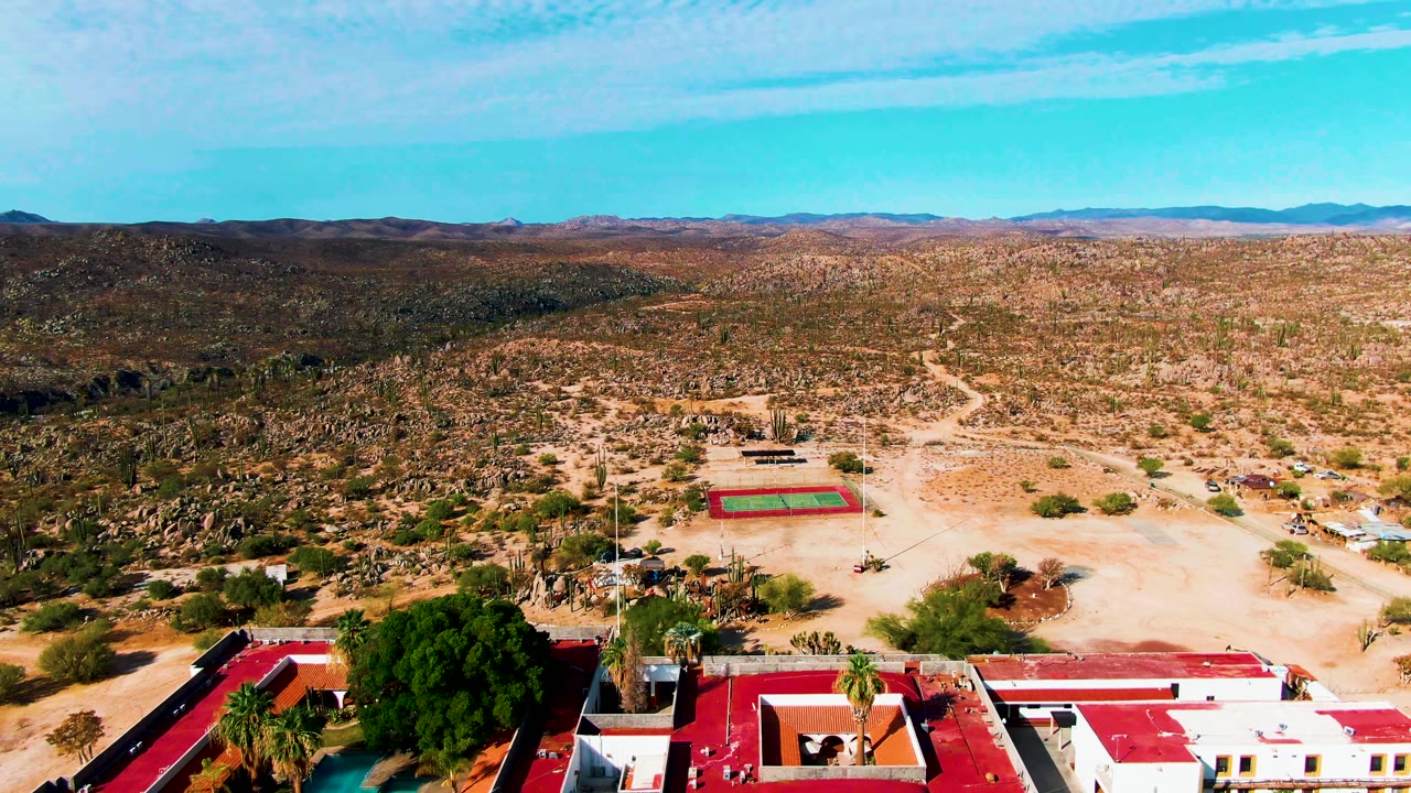 Drone frame over Catavina BC Mexico with red-roofed white buildings along the bottom edge, a tennis court farther out on sandy ground, desert vegetation, mountains, and no visible people.