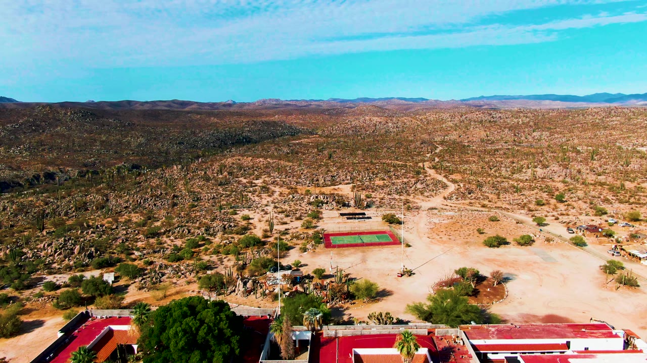 Catavina BC Mexico aerial frame where only the lower strip of red-roofed buildings remains, with the tennis court, pale dirt, cacti, open desert, mountain horizon, and no visible people.