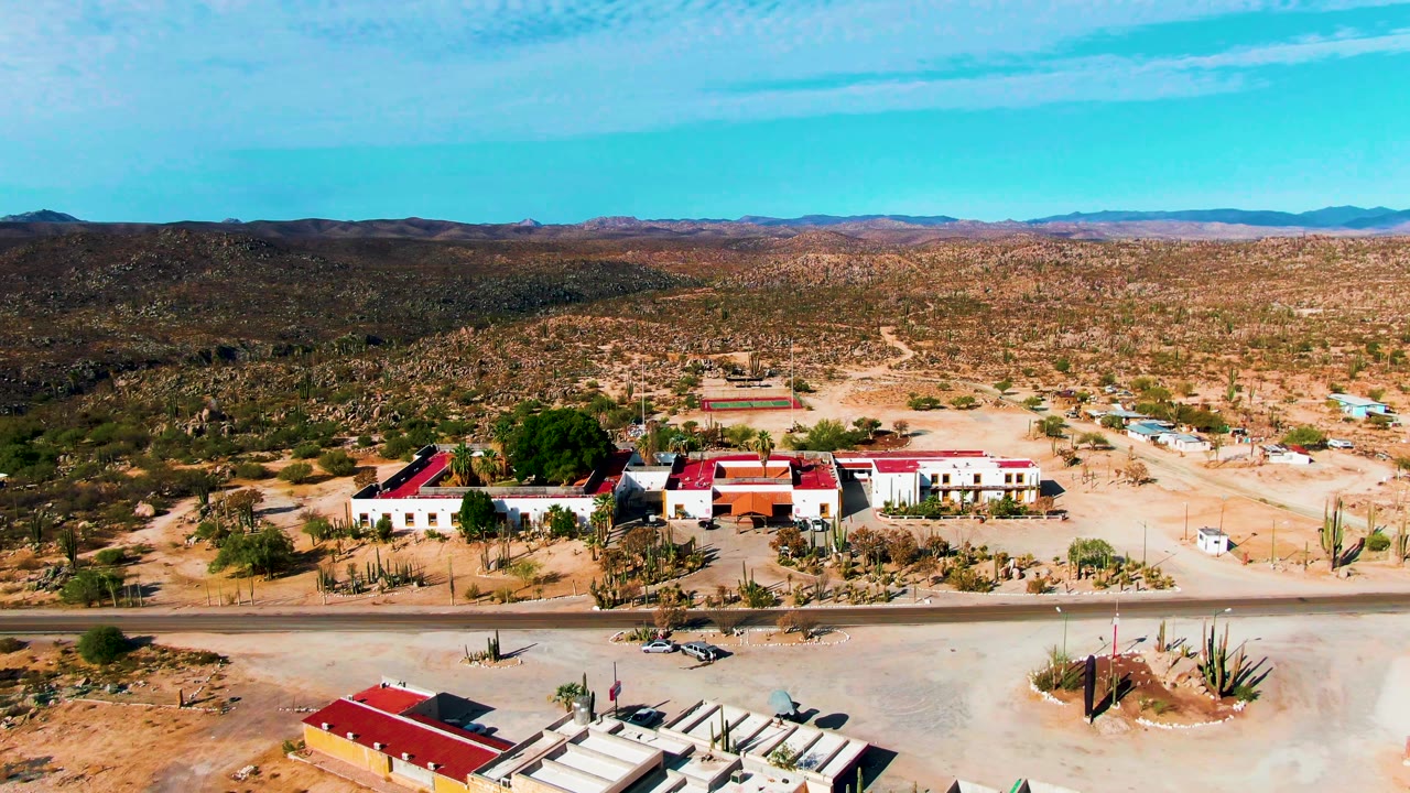 A high aerial frame over Catavina BC Mexico shows red-roofed buildings near a straight road, sandy ground, cacti, distant mountains, blue sky, and no visible people.