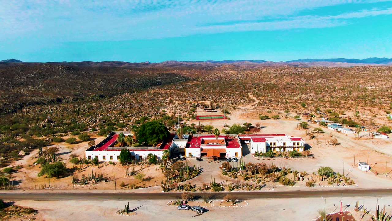 At 0:02, the aerial angle shows the red roofs and pale building walls more centered, with the road across the foreground, dry vegetation, hills, and no visible people.