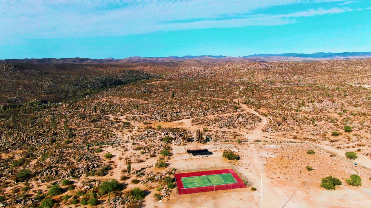 Aerial frame over Catavina BC Mexico showing a red and green tennis court near the bottom edge, a small shade structure, rocky desert, dirt tracks, distant hills, blue sky, and no visible people.