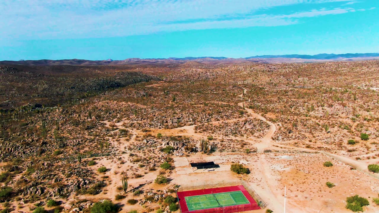 Aerial frame over Catavina BC Mexico with the tennis court partly lower in frame, a shade structure nearby, pale dirt roads, boulder-covered desert, scattered cacti, far hills, and no visible people.