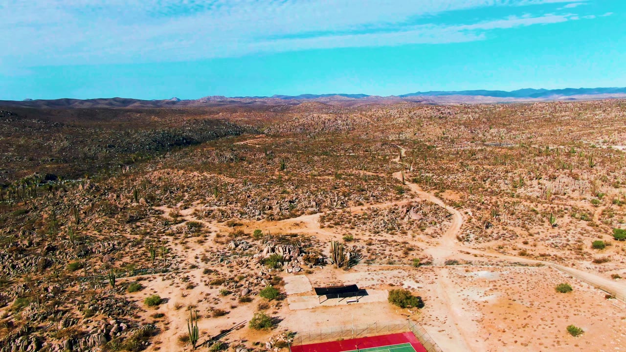 Aerial frame over Catavina BC Mexico showing the tennis court pushed toward the bottom edge, a nearby structure, winding dirt track, rocky desert vegetation, distant mountains, and no visible people.