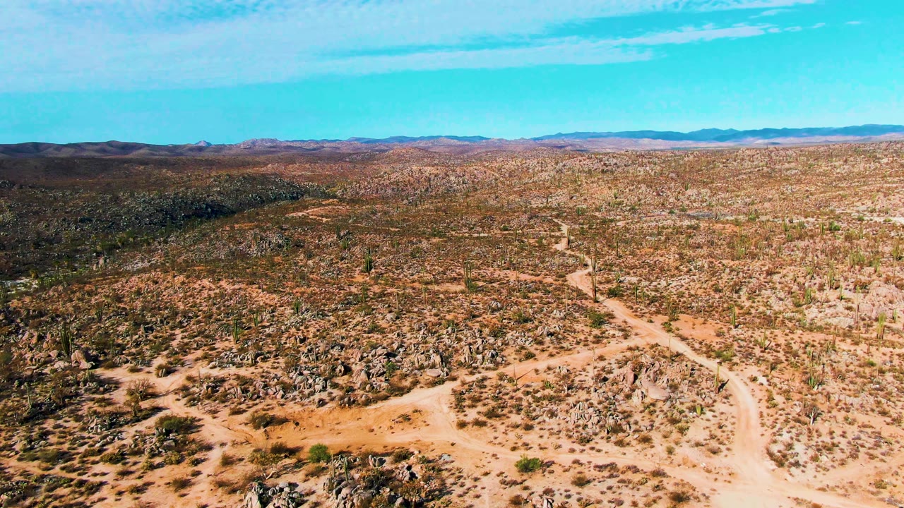 High drone-style frame over Catavina BC Mexico showing a pale dirt path bending through rocky desert, with cactus patches, dark hills on the left horizon, and no visible people.