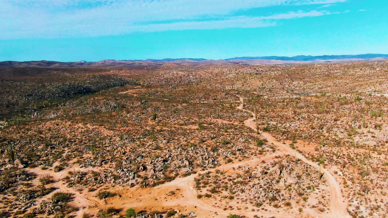 Aerial frame from Catavina BC Mexico with branching sandy tracks in the center, a rock-covered slope in the foreground, sparse desert vegetation, distant mountains, and no visible people.