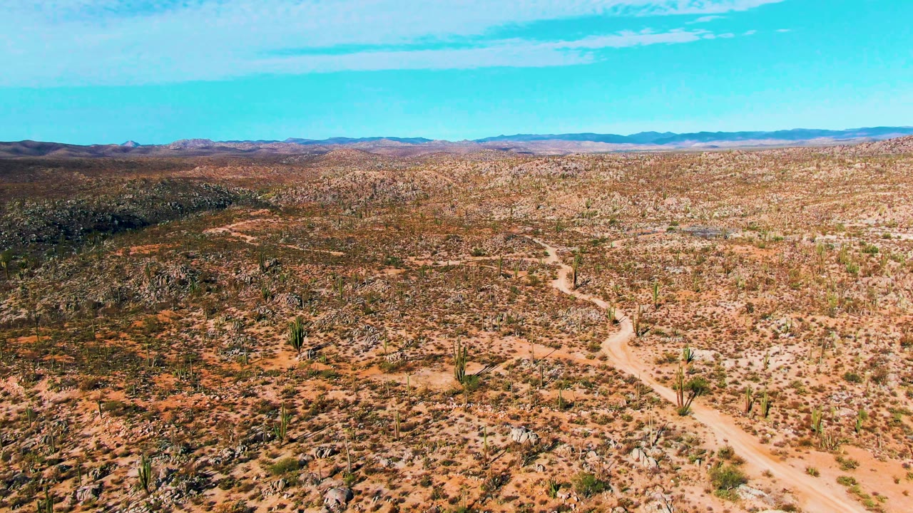 Early aerial frame over Catavina BC Mexico showing a pale dirt road curving up from the lower right through rocky desert with scattered cacti, distant mountains, blue sky, and no visible people.