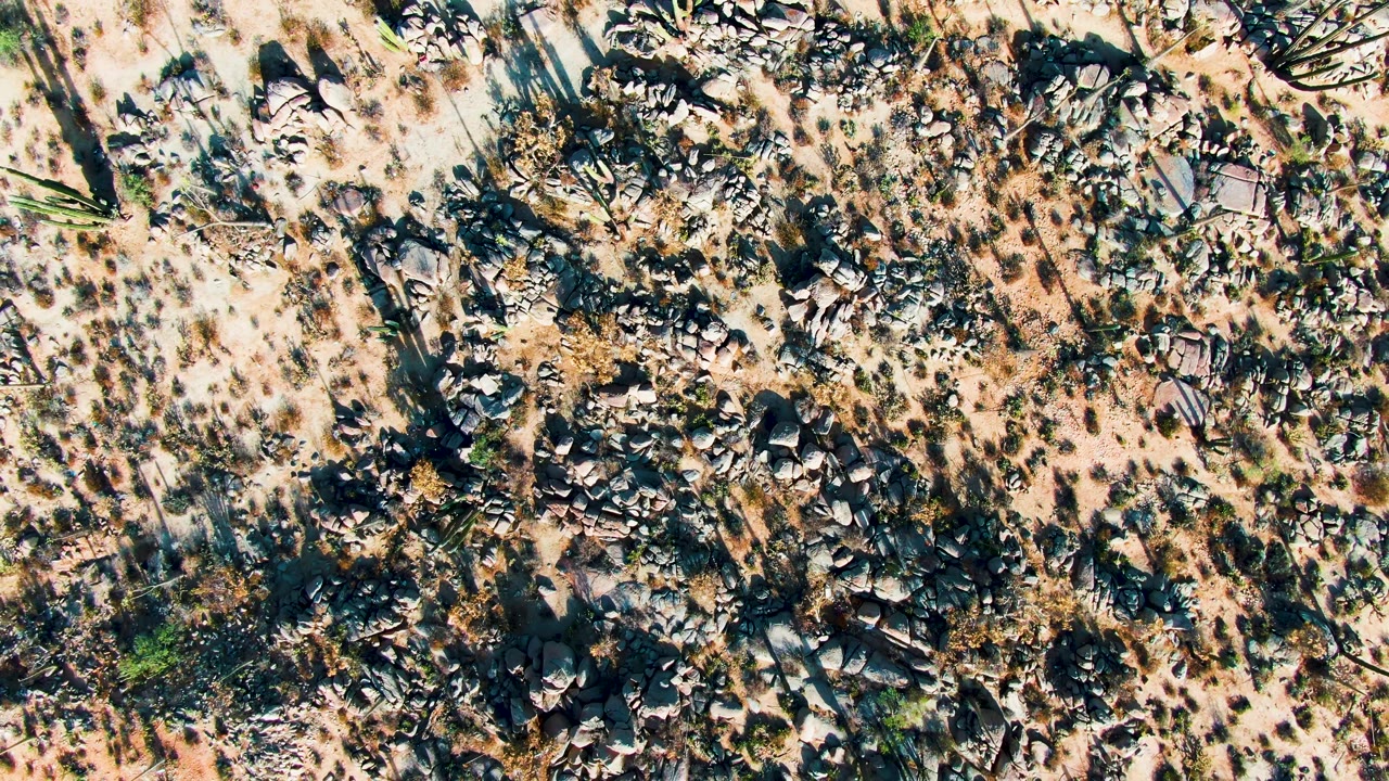 Aerial overhead view in Catavina BC Mexico showing denser dark rocks among beige sand, scattered brush, and no visible people.