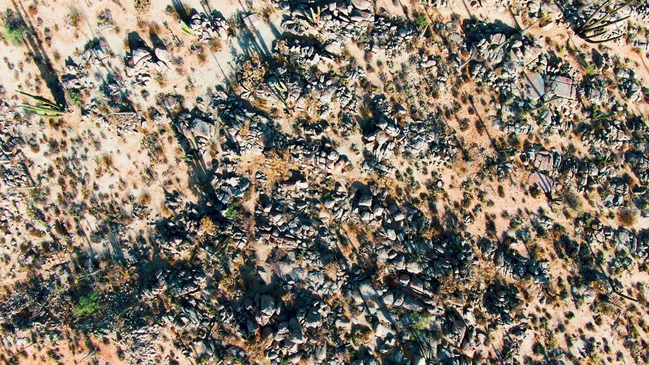 Overhead Catavina BC Mexico desert frame with mottled sand, broken rock clusters, isolated shrubs, and no visible people.