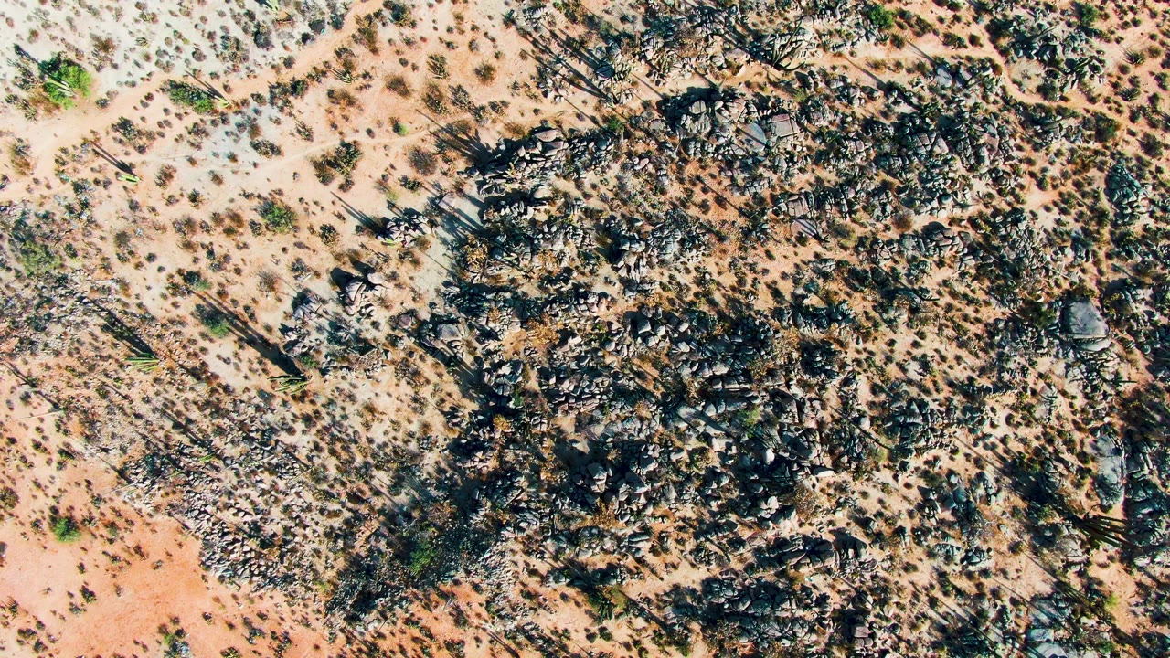 Aerial frame over Catavina BC Mexico showing a broad dark rock formation surrounded by pale sandy soil, small shrubs, and no visible people.