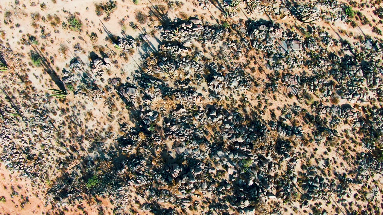 Aerial frame over Catavina BC Mexico desert terrain, showing a larger central band of dark rocks, scattered scrub, pale open sand, and no visible people.