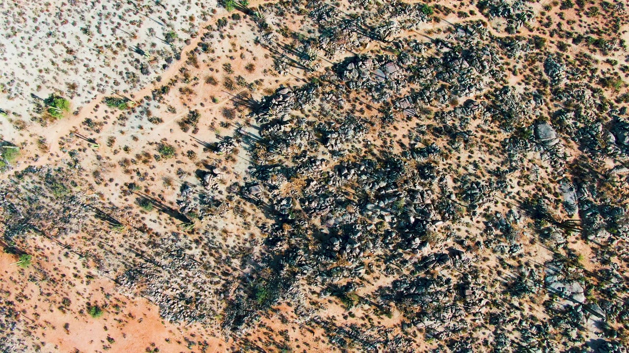 Overhead view of Catavina, Mexico desert terrain with dark rocky outcrops, pale sandy soil, scattered shrubs, and no visible people.