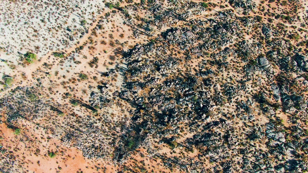 A Catavina, Mexico desert frame shows a dense dark rock cluster crossing sandy tan ground with small shrubs spread around it and no visible people.