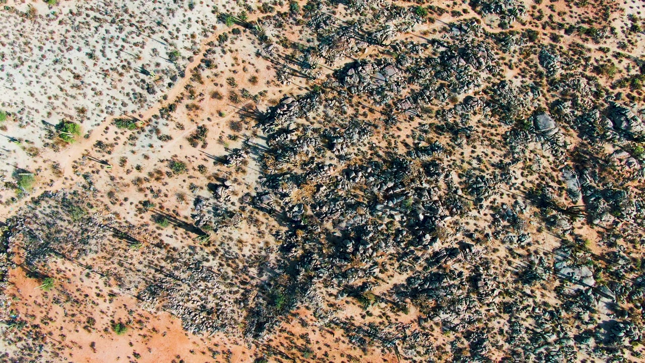 Overhead Catavina desert terrain fills the frame with black-brown rocks, beige sand, small greenish shrubs, and no visible people.