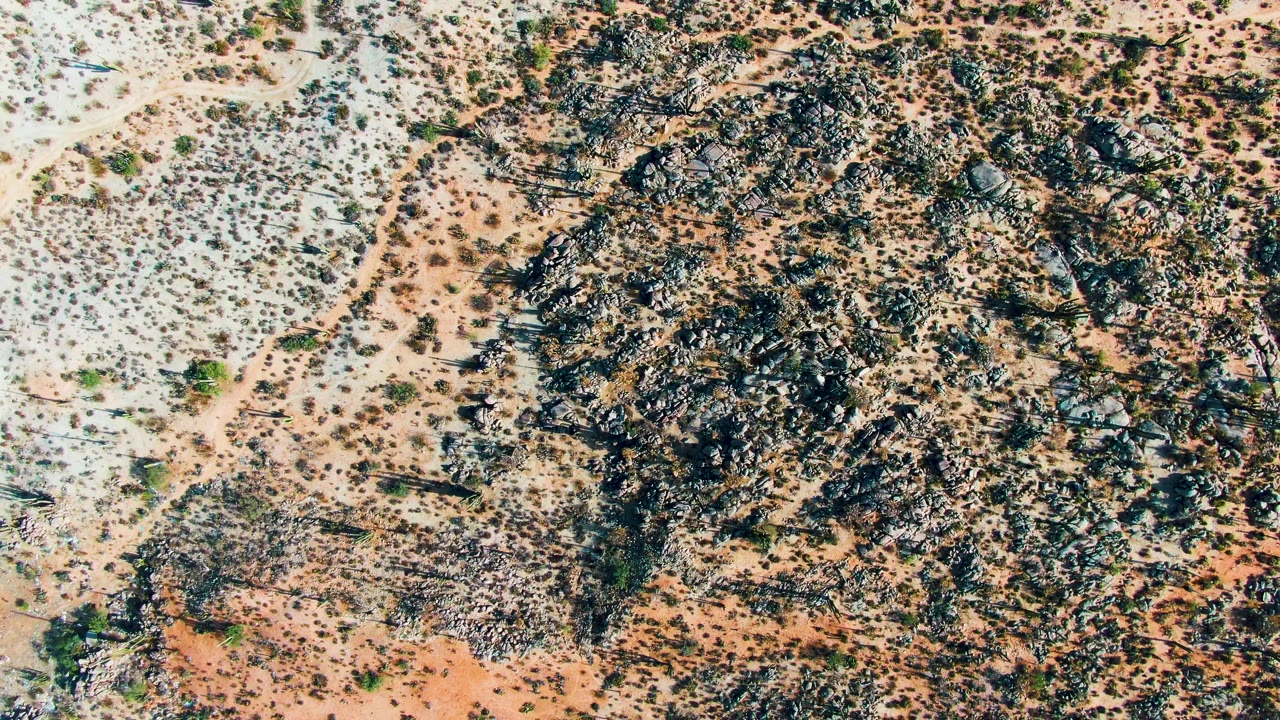 Aerial frame over Catavina, Mexico showing a denser band of dark rocks beside pale sandy desert and sparse green shrubs, with no visible people.