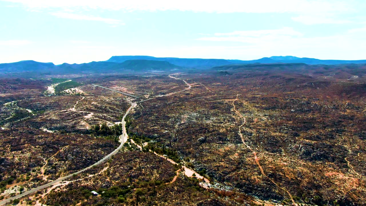 A high aerial frame over Catavina, BC Mexico shows a pale winding road crossing rocky brown desert toward distant flat-topped mountains, with no visible people.