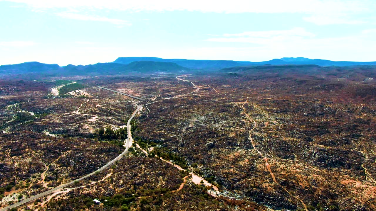 A broad aerial view in Catavina, BC Mexico shows the main road and smaller dirt tracks cutting through scrubby rock fields under a bright sky, with no visible people.