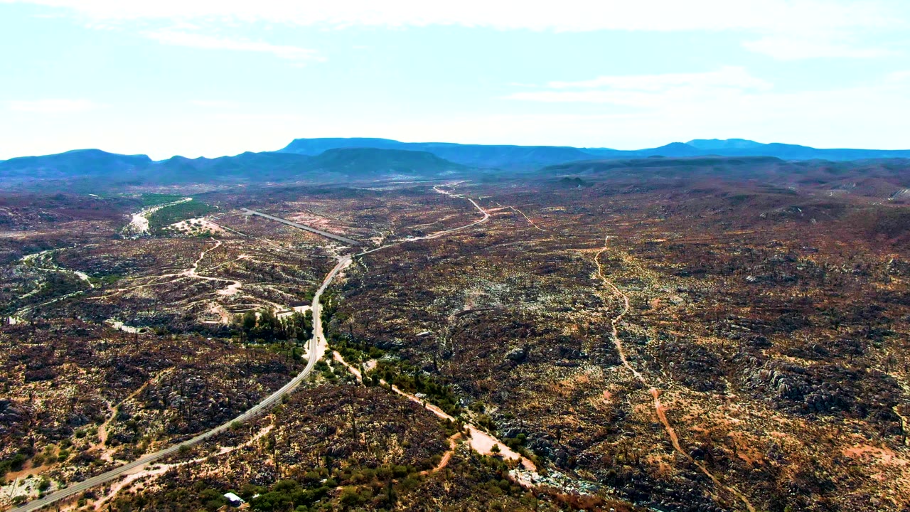 The aerial composition over Catavina, BC Mexico centers a curving pale road, mottled desert vegetation, exposed rocks, and distant blue mountains, with no visible people.