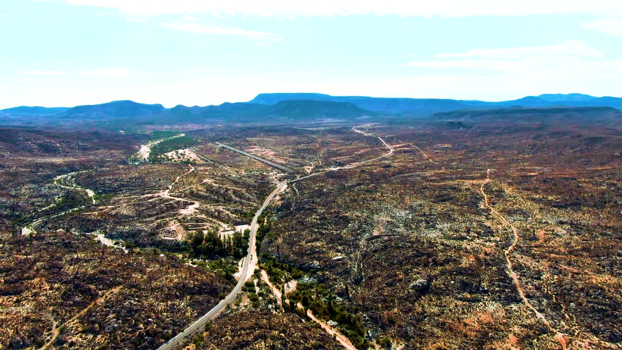 Opening aerial frame over Catavina BC Mexico shows a paved road and pale dirt tracks crossing brown-green desert hills toward distant mountain ridges, with no visible people.