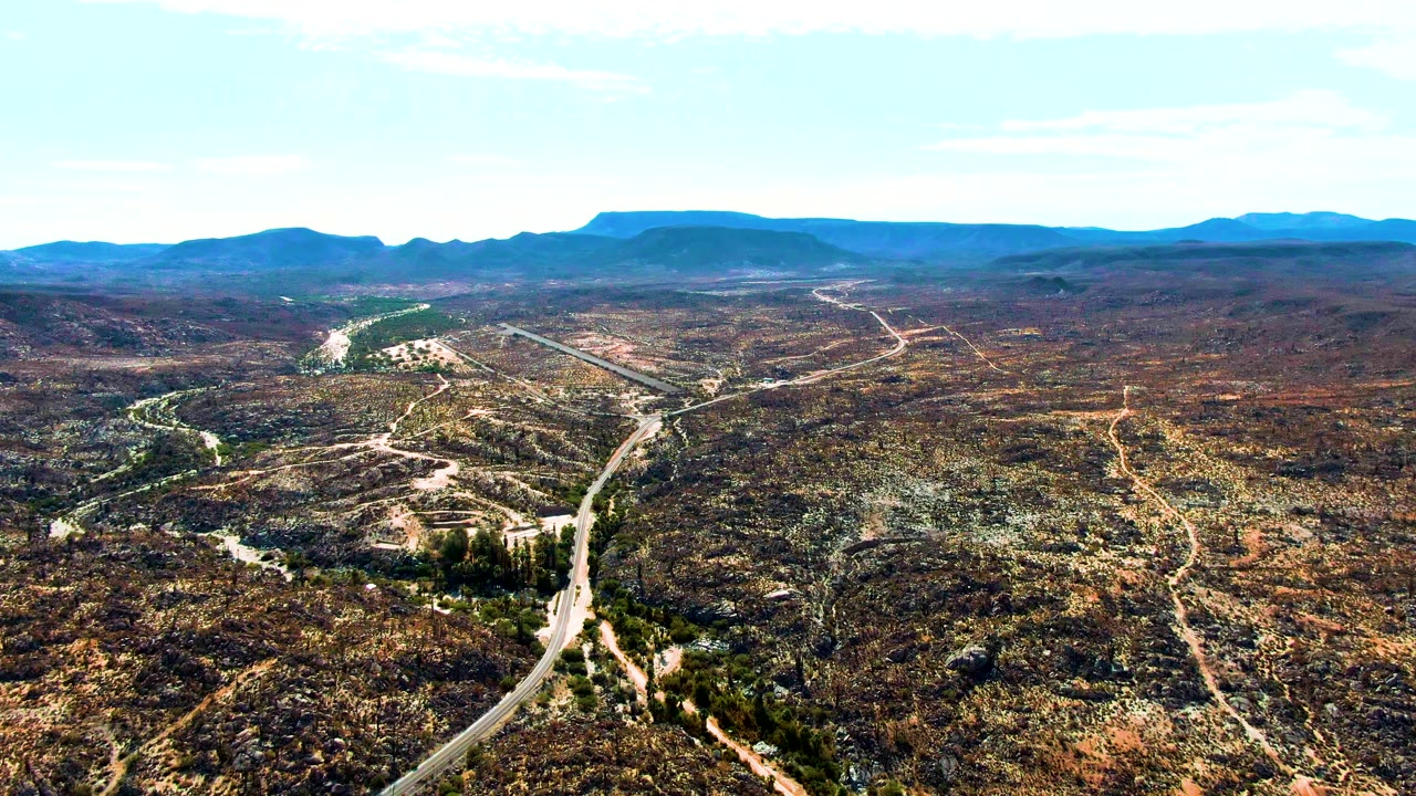 At 0:01, the Catavina BC Mexico aerial view holds broad desert scrub, a curving road in the lower frame, a greener wash left of center, and blue ridges on the horizon, with no visible people.
