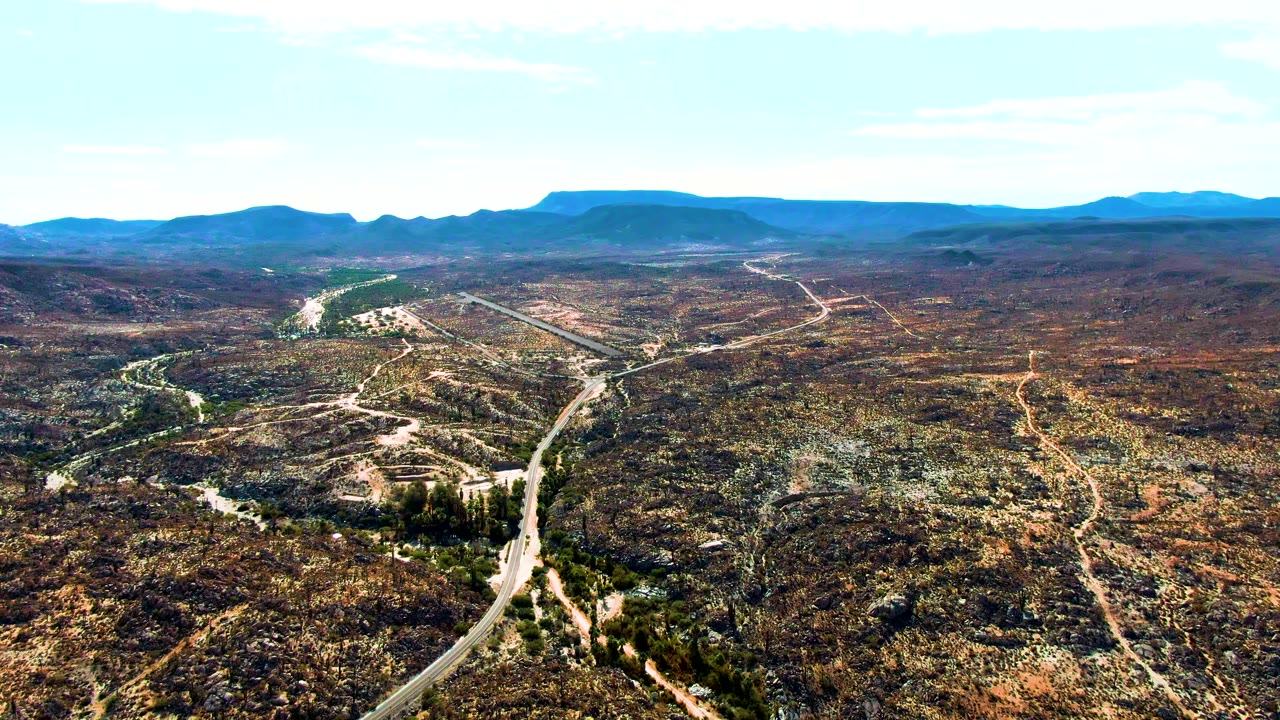 At 0:02, a high aerial view of Catavina BC Mexico shows the main road running upward from the lower edge, smaller tracks branching through arid hills, and flat-topped distant mountains, with no visible people.