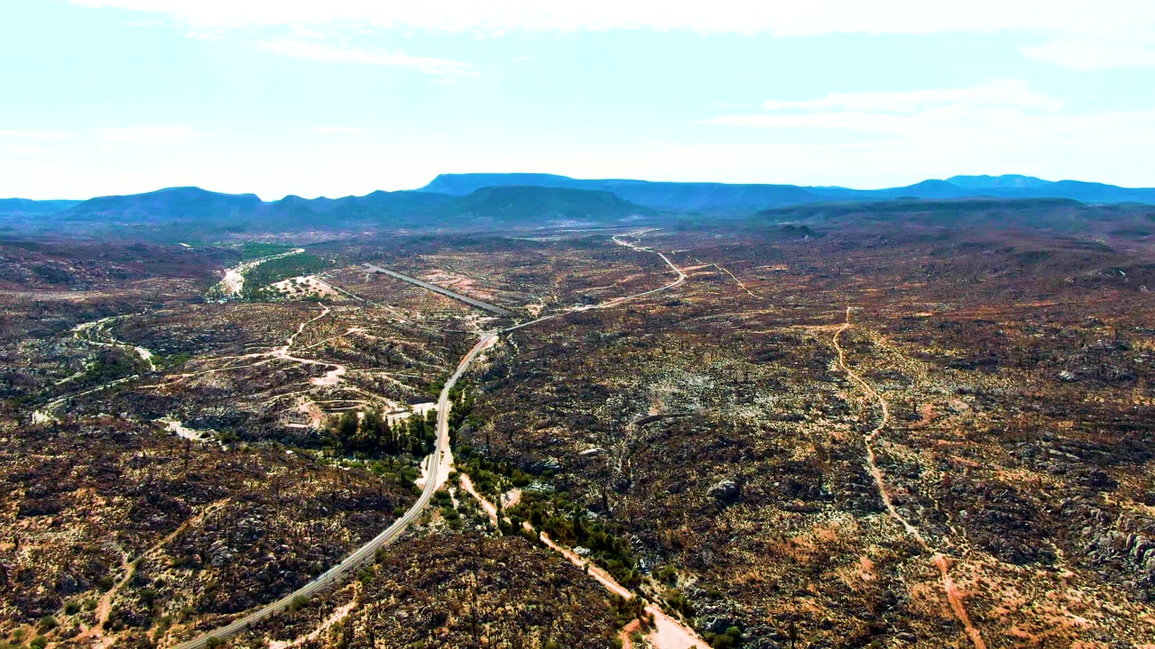 High aerial frame of Catavina BC Mexico with the road curving from the lower edge toward the midground, scrub vegetation on both sides, far hills, and no visible people.