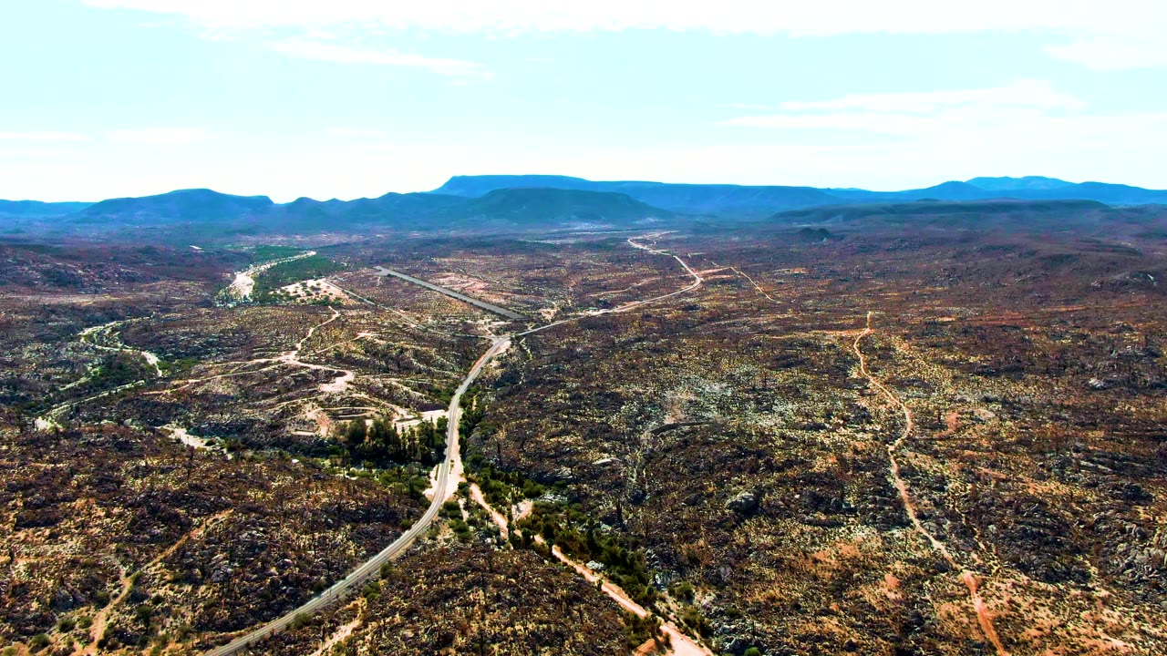 Wide drone perspective over Catavina BC Mexico showing the winding road threading through arid brown and green terrain beneath a pale blue sky, with no visible people.