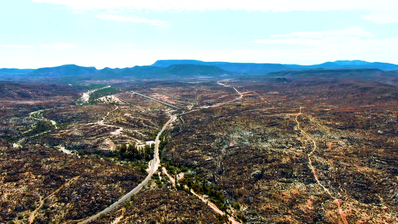 Drone view in Catavina BC Mexico where the light road crosses dry rolling terrain, small tracks branch through scrub, distant mountains sit along the horizon, and no visible people appear.