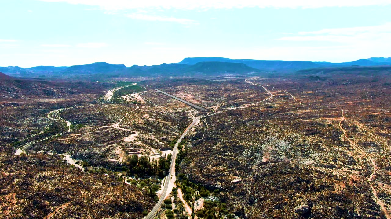 Wide aerial frame over Catavina BC, Mexico, showing a pale winding road through arid scrub hills, distant blue mountains, bright clear sky, and no visible people.