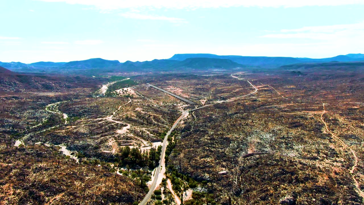 A high aerial view in Catavina BC, Mexico, with the road running from the lower frame into dry hills, pale dirt tracks across the terrain, distant ridges, and no visible people.