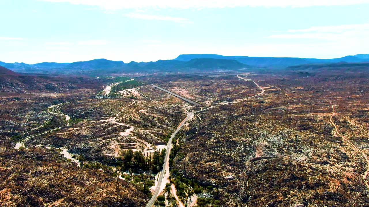 Aerial frame from Catavina BC, Mexico, showing a small vehicle on a pale road, scrub-covered hills across the foreground and midground, blue mountain ridges beyond, and no visible people.