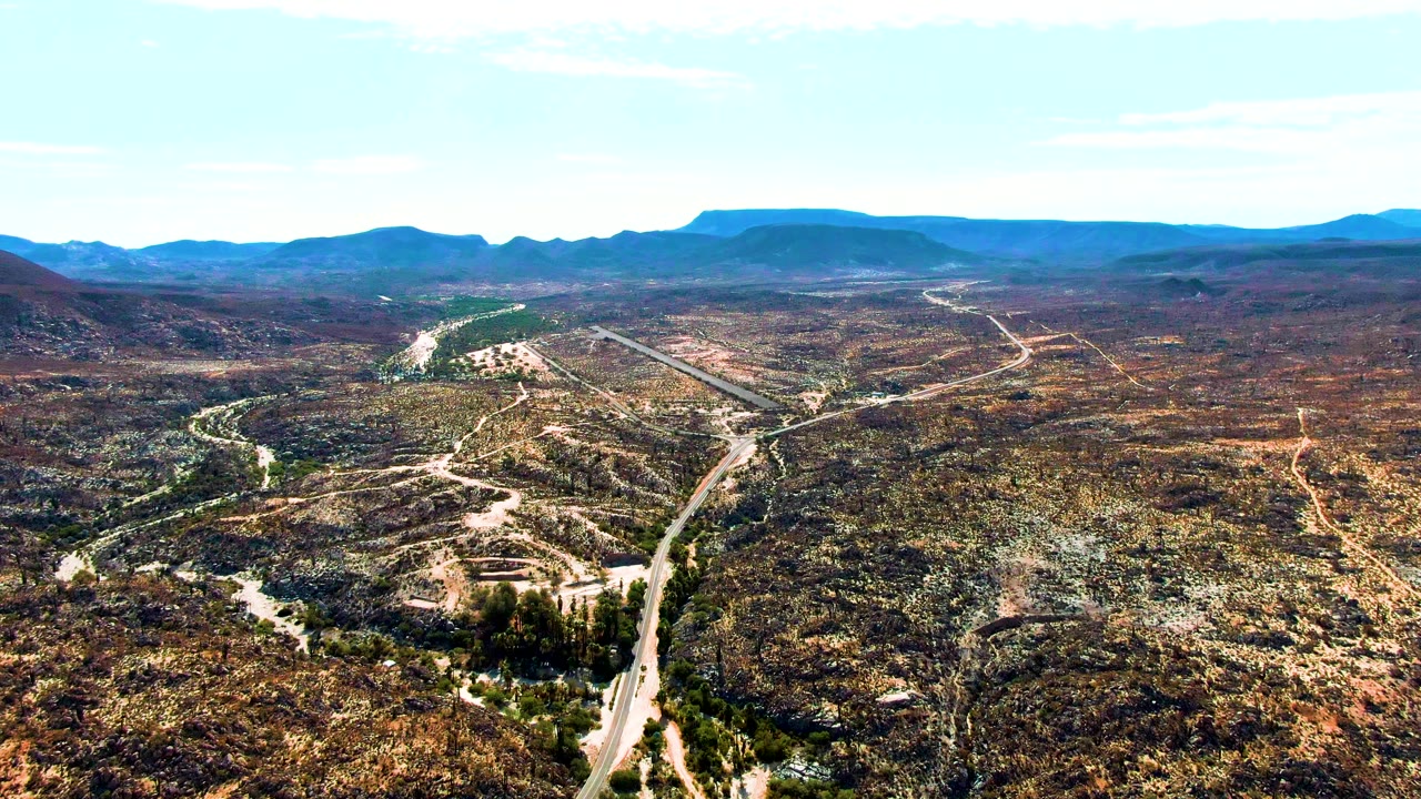 Wide aerial view over Catavina BC, Mexico, with the road curving up from the lower edge through mottled brown and green scrub hills, distant mountains, clear sky, and no visible people.