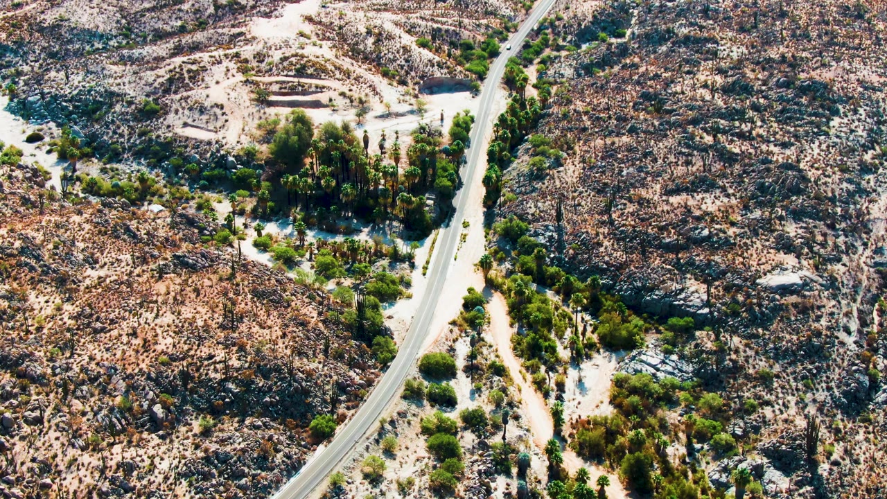 Aerial frame from Catavina, Mexico showing a paved road bending through rocky desert hills with green vegetation near sandy ground, no visible people.