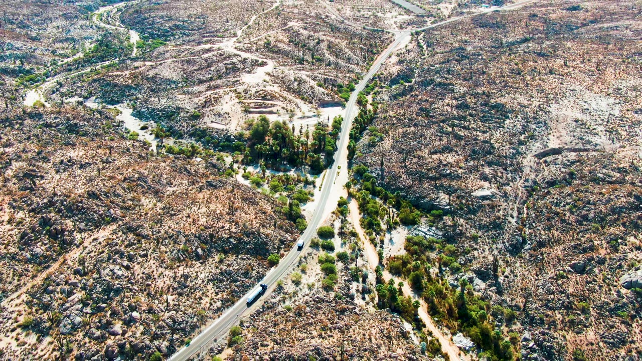 Aerial frame over Catavina BC, Mexico showing the winding road cutting past a green wash area, the vehicle on the pavement, dry rocky hills, and no visible people.